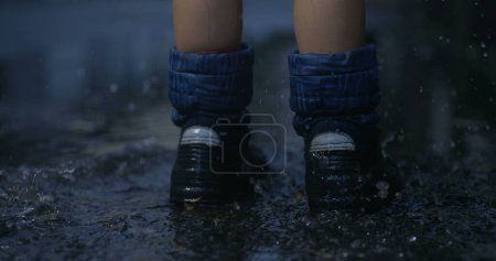 Téléchargez les photos : Gros plan enfant portant des bottes de pluie sautant dans la flaque d'eau capturée avec une caméra haute vitesse éclaboussant des gouttelettes liquides dans l'air - en image libre de droit