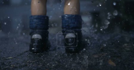 Foto de Primer plano del niño con botas de lluvia salpicando en charco con captura de alta velocidad, los pies del niño en ropa de lluvia saltando en el charco de agua causando salpicaduras de gotas - Imagen libre de derechos