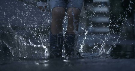 Foto de Un niño excitado con botas de lluvia salta al charco de agua en cámara lenta capturando gotas en el aire con una cámara de alta velocidad, temporada de lluvias - Imagen libre de derechos
