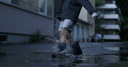 Foto de Nostálgica escena de niño pateando charco de agua en callejón de la calle divirtiéndose por sí mismo filmado en cámara de alta velocidad - Imagen libre de derechos