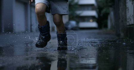 Foto de Niño despreocupado corriendo con botas de lluvia en charcos de agua en la calle durante la noche en ultra cámara lenta 800 fps salpicando gotas en el aire - Imagen libre de derechos