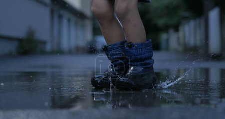 Téléchargez les photos : Enfant portant des bottes de pluie marchant sur la flaque d'eau créant des éclaboussures volant dans les airs - en image libre de droit