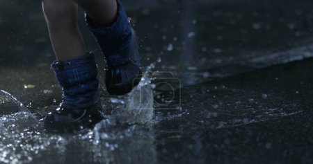 Téléchargez les photos : Enfant marchant dans la flaque d'eau au ralenti super portant des bottes de pluie éclaboussant des gouttelettes liquides partout capturées avec une caméra haute vitesse en 800 ips - en image libre de droit