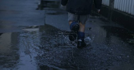 Téléchargez les photos : Enfant sprinter joyeusement dans des bottes de pluie dans la flaque d'eau avec éclaboussure au ralenti super - en image libre de droit
