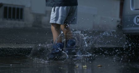 Téléchargez les photos : Enfant éclaboussant de l'eau partout tombant dans la flaque dans la rue portant des bottes de pluie dans une caméra à grande vitesse à 800 ips - en image libre de droit