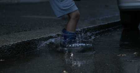 Téléchargez les photos : Enfant sautant dans la flaque d'eau dans la rue avec des bottes de pluie au ralenti à 800 ips capturées à grande vitesse, éclaboussant l'eau partout - en image libre de droit
