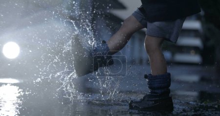 Foto de Niño despreocupado jugando con charco de agua en la calle patadas y salpicaduras gotitas en el aire en cámara súper lenta capturado con cámara de alta velocidad - Imagen libre de derechos