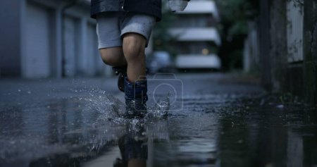 Téléchargez les photos : Enfant insouciant courant avec des bottes de pluie sur les flaques d'eau dans la rue le soir en ultra-lent 800 ips éclaboussant des gouttelettes dans l'air - en image libre de droit