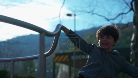 Téléchargez les photos : Petit garçon tournant délicieusement sur le rond-point du parc, enfant énergique s'amusant de manière insouciante. enfant jouant joyeusement sur le carrousel à l'aire de jeux - en image libre de droit
