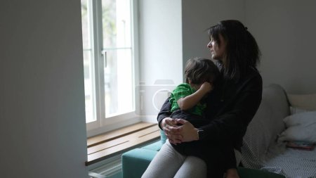 Téléchargez les photos : Mère consolant enfant blessé par la fenêtre à la maison. Candid tendre moment de pensif maman réconfortante petit garçon - en image libre de droit