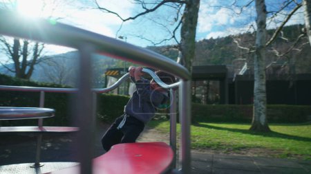 Téléchargez les photos : Petit garçon tourbillonnant joyeusement sur le carrousel de l'aire de jeux, enfant profitant d'un tour sur l'équipement rond-point du parc - en image libre de droit