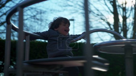 Téléchargez les photos : Petit garçon tournant délicieusement sur le rond-point du parc, enfant énergique s'amusant de manière insouciante. enfant jouant joyeusement sur le carrousel à l'aire de jeux - en image libre de droit