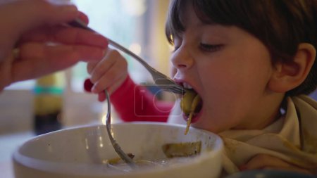 Foto de Madre alimentando a su hijo con espaguetis comida de pasta en un tazón cubierto con servilleta en el cuello. Niño disfruta de la comida italiana - Imagen libre de derechos