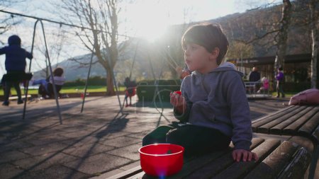Téléchargez les photos : Petit garçon assis au parc en train de grignoter tout en observant son environnement. L'enfant déguste une collation à l'aire de jeux au soleil pendant la saison automnale - en image libre de droit