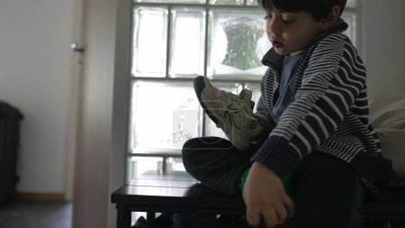 Téléchargez les photos : Petit garçon essayant de mettre chaussure par porte d'appartement devant. enfant lutte avec des chaussettes et des chaussures. enfant n'aime pas la sensation, l'irritation quand les vêtements - en image libre de droit