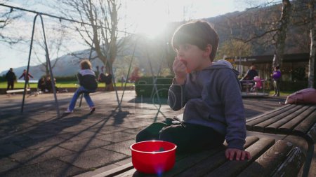 Photo for Small boy seated at park snacking while observing his surroundings. Child enjoys food snack at playground in the sunlight flare during fall season - Royalty Free Image