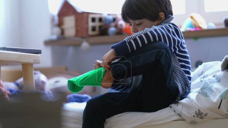 Téléchargez les photos : Enfant portant une chaussette assise au chevet du lit. Petit garçon s'habillant lui-même. Le gamin se prépare à commencer la journée. - en image libre de droit