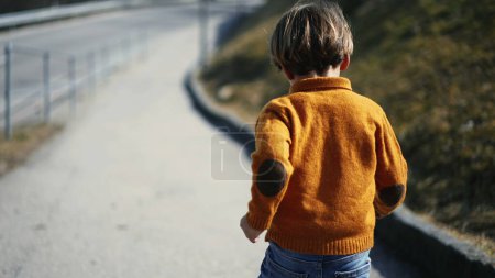 Photo for Rear view of an eager young boy, dashing outdoors amidst the fall ambiance. Clad in a yellow pullover, jeans, and boots, the child revels in the joy of an autumnal sprint - Royalty Free Image