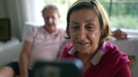 Photo for Happy grandmother laughing and smiling while interacting with relatives in video call. Long-distance communication between grandparents seated on couch speaking with family - Royalty Free Image