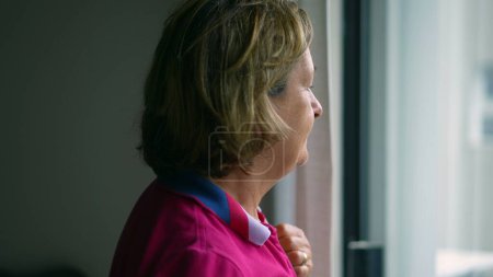 Photo for Contemplative retired senior woman standing by window gazing out at view, profile close-up of elderly lady in mental reflection - Royalty Free Image
