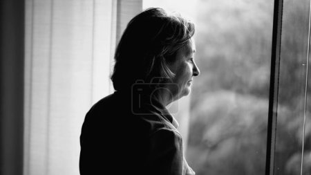 Foto de Mujer anciana reflexionando por la ventana en el día lluvioso en la escena monocromática, en blanco y negro de la señora jubilada perdida en el pensamiento viendo la lluvia - Imagen libre de derechos