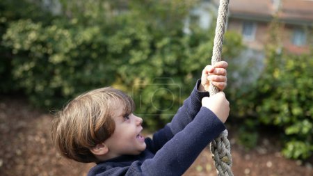Foto de El niño se divierte en el parque infantil sosteniéndose en la cuerda y deslizándose hacia abajo durante la temporada de otoño. Niño feliz disfrutando de momentos de infancia - Imagen libre de derechos