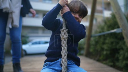 Photo for Carefree small boy sliding down wire rope gripping firmly while descending wire attached between two trees during autumn fall day at public park. Child having fun - Royalty Free Image
