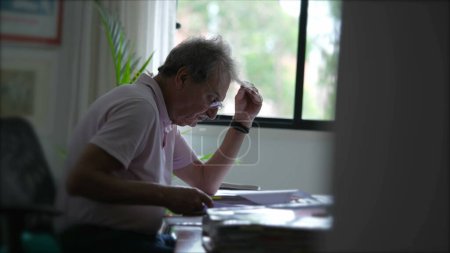 Photo for Candid senior man working at home inspecting documents concentrated reading paperwork. Older person in office desk - Royalty Free Image