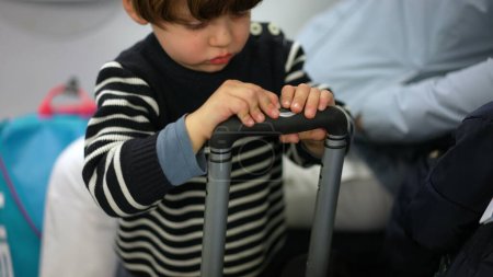 Foto de Niño aburrido juega con el asa de la maleta mientras viaja con la familia, curioso niño pequeño que pasa el tiempo con el objeto de equipaje - Imagen libre de derechos