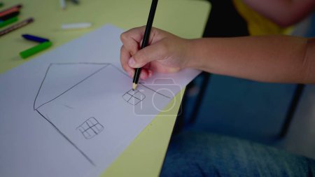 Photo for Close-up child hand drawing a traditional house, two windows and a front door with pencil on paper, creativity play time childhood concept - Royalty Free Image