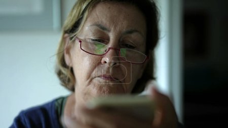 Téléchargez les photos : Gros plan sur le visage d'une femme âgée lisant du contenu en ligne avec un appareil smartphone. Une dame caucasienne âgée regardant l'écran du téléphone avec des lunettes - en image libre de droit