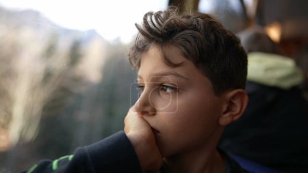 Photo for Melancholic young boy sitting by train window looking at view with hand in chin, thoughtful pensive expression of pre-teen child traveling and daydreaming - Royalty Free Image