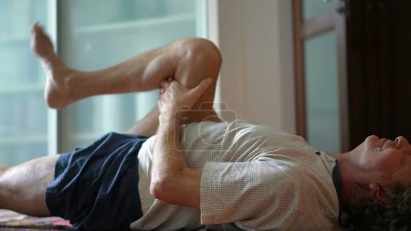 Foto de Anciano haciendo estiramientos de la pierna en su espalda, cuidando de su salud en la vejez. Hombre caucásico retirado enfocándose en ejercicios de piernas matutinas - Imagen libre de derechos