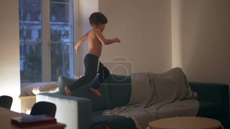 Téléchargez les photos : Enfant franc jouant la nuit sur le canapé, courant énergiquement le soir en s'amusant seul le soir - en image libre de droit