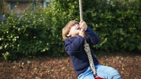 Foto de Niño pequeño alegremente deslizándose por la cuerda de alambre entre los árboles en Autumn Park - Imagen libre de derechos