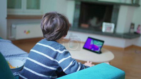 Téléchargez les photos : Enfant regardant bande dessinée à la maison assis sur le canapé, petit garçon absorbé par le contenu de divertissement sur l'écran de l'appareil tablette - en image libre de droit