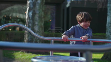 Téléchargez les photos : Enfant jouant au manège de l'aire de jeux pendant la saison automnale. Petit garçon tournant en rond tout en tenant sur barre métallique obtenir l'exercice et l'activité de plein air - en image libre de droit