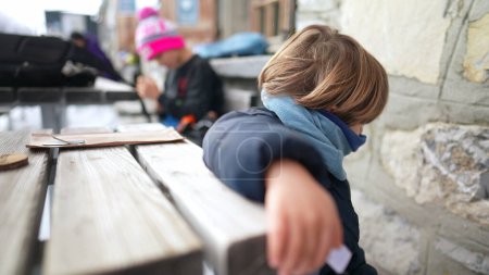 Photo for Little boy wearing scarf and jacket during winter season sitting by table outside waiting. Child wears warm clothes in cold day - Royalty Free Image