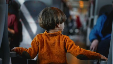 Téléchargez les photos : Un garçon vêtu d'un pull jaune se promène dans l'allée du train, saisissant les poignées de siège pour le soutenir. Le jeune voyageur passe son temps à errer à bord du wagon - en image libre de droit