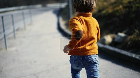 Téléchargez les photos : Dos d'un jeune garçon insouciant courant dehors portant un pull jaune et des bottes pendant la journée d'automne. Sprint enfant, engagé dans l'exercice quotidien - en image libre de droit