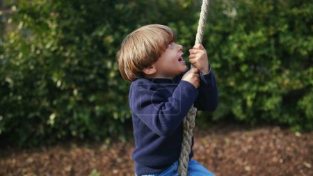 Téléchargez les photos : Petit garçon glissant joyeusement le long de la corde métallique entre les arbres dans le parc d'automne - en image libre de droit
