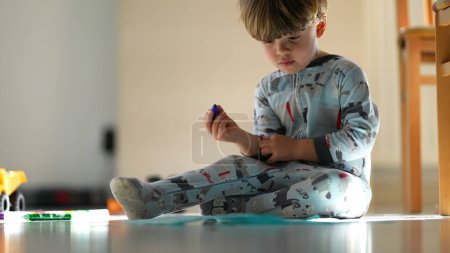 Téléchargez les photos : Enfant portant un pyjama tôt le matin assis sur le dessin au sol sur papier avec un stylo à coloration bleue - en image libre de droit