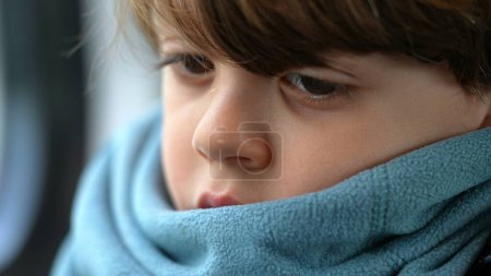 Téléchargez les photos : Visage d'enfant pensif en gros plan dans une émotion profonde et réfléchie portant une écharpe. Un petit garçon caucasien perdu dans la pensée - en image libre de droit