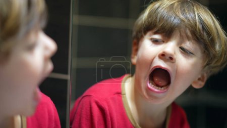 Téléchargez les photos : Enfant regarde miroir de salle de bain vérifier la bouche et les dents, petit garçon ouvrant la bouche en regardant son reflet - en image libre de droit
