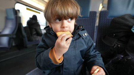 Foto de Niño merendando galletas de mantequilla mientras viaja en tren. Niño vistiendo ropa de invierno comiendo bocadillos mientras mira a la ventana con expresión pensativa - Imagen libre de derechos