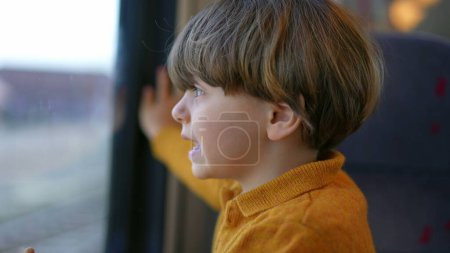 Téléchargez les photos : Gros plan visage d'enfant regardant depuis la fenêtre du train avec une expression curieuse. Un petit garçon caucasien voyageant - en image libre de droit