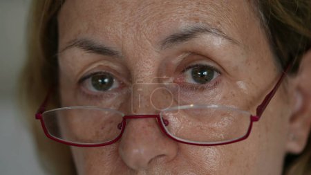 Téléchargez les photos : Senior lady fermer les yeux et le visage en regardant la caméra avec des lunettes de lecture. Personne âgée caucasienne dans les années 70 regardant dans une expression neutre - en image libre de droit