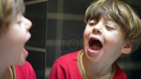 Téléchargez les photos : Enfant regarde miroir de salle de bain vérifier la bouche et les dents, petit garçon ouvrant la bouche en regardant son reflet - en image libre de droit
