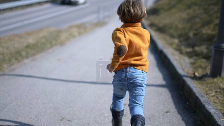 Téléchargez les photos : Dos de petit garçon excité sprinter à l'extérieur pendant la journée d'automne, Enfant portant pull jaune, jeans et bottes court - en image libre de droit