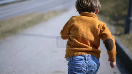 Foto de Espalda de niño pequeño excitado corriendo al aire libre durante el día de otoño, niño usando jersey amarillo, jeans y botas corre - Imagen libre de derechos